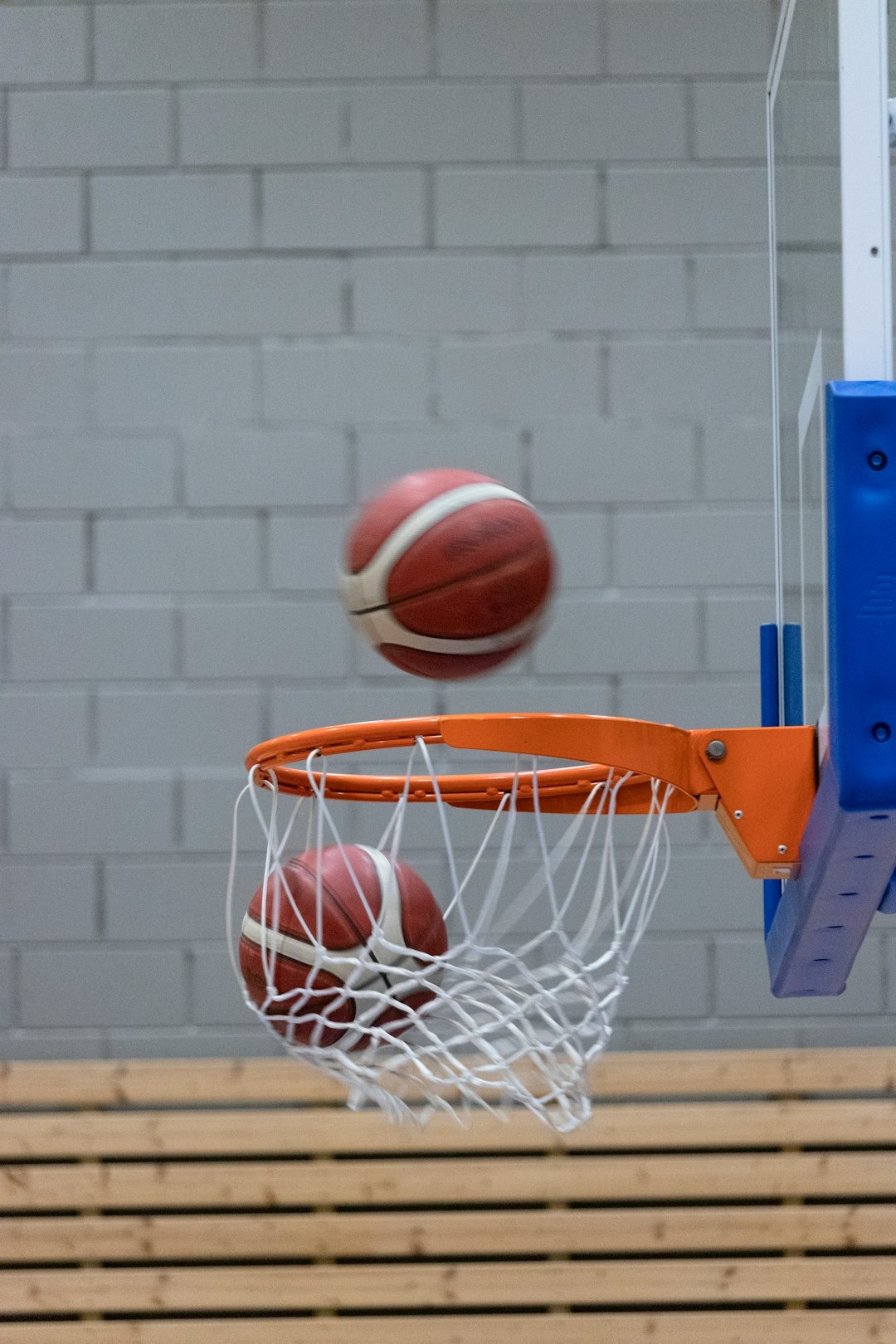 Two basketballs pass through a hoop on an indoor court.